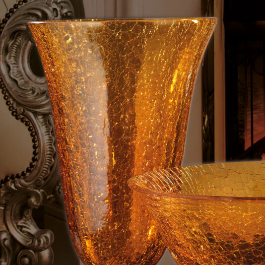 Luxury Amber Glass Vase And Bowl An image showcasing the Luxury Amber Glass Vase and Bowl set, featuring two elegant, golden glass pieces with a crackled texture. The taller vase is prominently displayed at the forefront, while a shorter bowl with a similar style is partially visible beside it. The background features an ornate decorative element, enhancing the luxurious amber glass vibe.