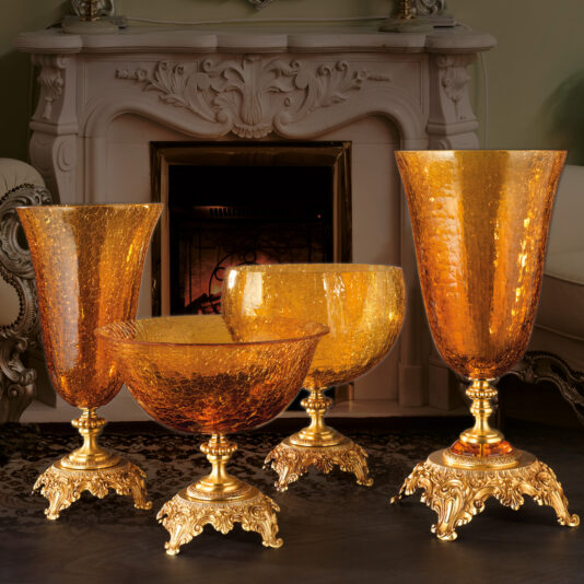 A luxurious set of four Luxury Amber Glass Vase And Bowl pieces with intricate gold bases, meticulously arranged in front of an elegantly carved white marble fireplace. The room exudes opulence with its detailed furnishings and ambient lighting.