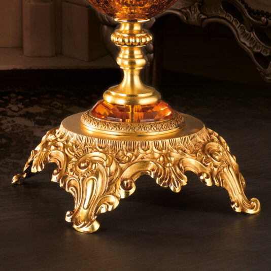 Luxury Amber Glass Vase And Bowl Close-up of the ornate, gold-colored base of a decorative item, possibly a lamp or trophy, resting on a dark surface. The base features intricate carvings and a reflective amber gem-like element, exuding the luxurious aesthetic reminiscent of the Luxury Amber Glass Vase And Bowl.