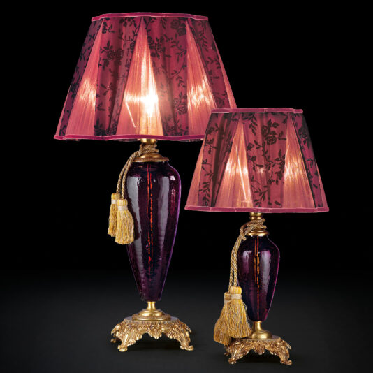 A pair of Luxury Amethyst Glass Table Lamps, each featuring a purple base adorned with gold accents and tassels, comes with matching magenta lampshades decorated in floral patterns. One lamp stands slightly taller than its counterpart, creating a visually appealing duo against the black background.