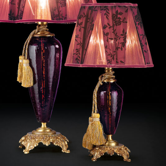 Luxury Amethyst Glass Table Lamp The Luxury Amethyst Glass Table Lamp comes as a set of two, featuring stunning amethyst glass bases with gold accents. Each lamp is topped with a wide, pleated fabric shade embellished with black floral designs and finished with a gold tassel. The lamps are supported by intricate gold bases that boast a detailed design, exuding elegance and sophistication.