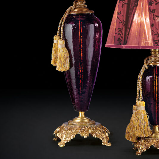 Luxury Amethyst Glass Table Lamp A pair of Luxury Amethyst Glass Table Lamps featuring deep purple glass bodies with ornate golden bases and decorative tassels. These exquisite lamps are adorned with elaborate gold detailing at the base and a matching finial. One lamp's shade is partially visible, showcasing intricate patterns against a black background.