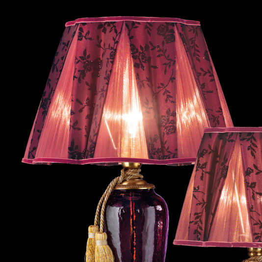 Luxury Amethyst Glass Table Lamp The Luxury Amethyst Glass Table Lamp showcases a stunning dark red and black floral-patterned shade. It is complemented by a shiny, dark base adorned with gold accents, including an elegant golden rope with tassels. The warm glow from the lamp creates a cozy atmosphere, and another similar lampshade made of amethyst glass can be seen partially in view.