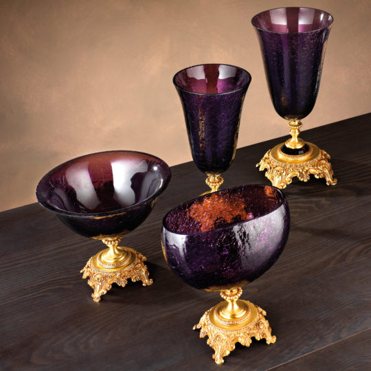 A set of four elegant items, part of the Luxury Amethyst Glass Vase And Bowl collection, is displayed on a dark wooden surface. Two pieces are tall and narrow like vases, while the other two resemble bowls with wide openings. The exquisite purple glass paired with intricate gold detailing creates a luxurious aesthetic.