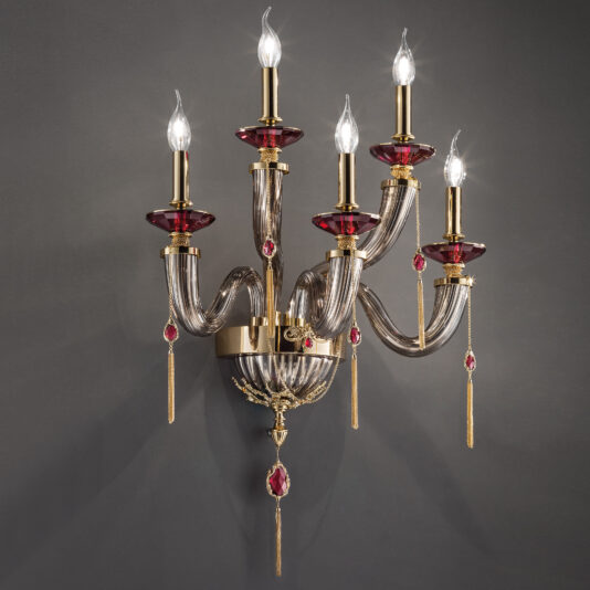 A sophisticated gold wall-mounted candelabra with five gracefully curving arms, each hosting a candle-shaped light bulb. The arms are adorned with clear, twisted glass elements and red, gem-like decorations, reminiscent of the Mirrored Glass Candle Style Wall Light With Swarovski® Crystals. This exquisite piece radiates opulence and classic elegance.