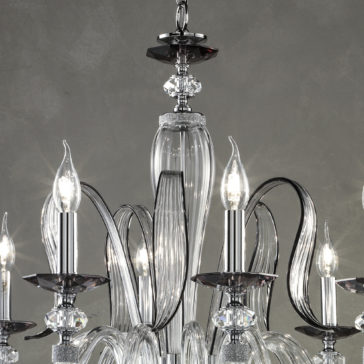 A close-up of the Modern Chandelier With Swarovski® Crystals showcases its elegant, modern design with intricate, curved glass arms and multiple candle-shaped light bulbs. Adorned with sparkling Swarovski® crystals, the chandelier features clear glass elements and a sleek silver finish against a dark gray background.