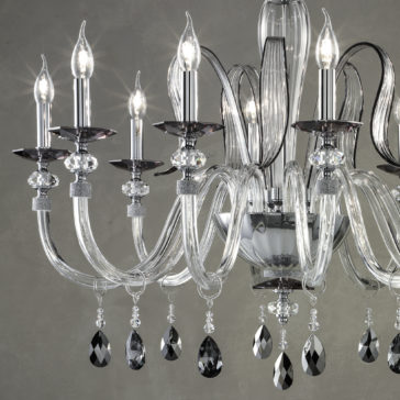 The Modern Chandelier With Swarovski® Crystals boasts luxurious, intricately designed glass arms and five candle-style light fixtures, each holding a glowing bulb. Hanging teardrop-shaped black and clear crystal ornaments enhance the elegance of the design, all set against a neutral backdrop.