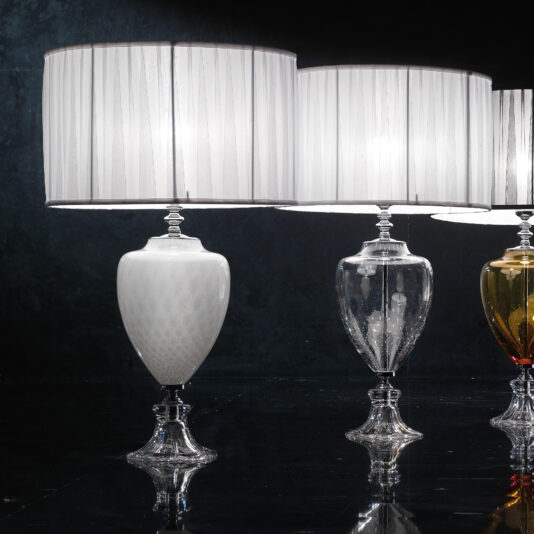 Modern Coloured Glass Table Lamps The Modern Coloured Glass Table Lamps feature three exquisite and elegant designs, each with a pleated white shade and an ornate base. The bases are artistically intricate, offered in white, clear, and amber variations. These lamps emit a beautiful, soft glow against a dark background.