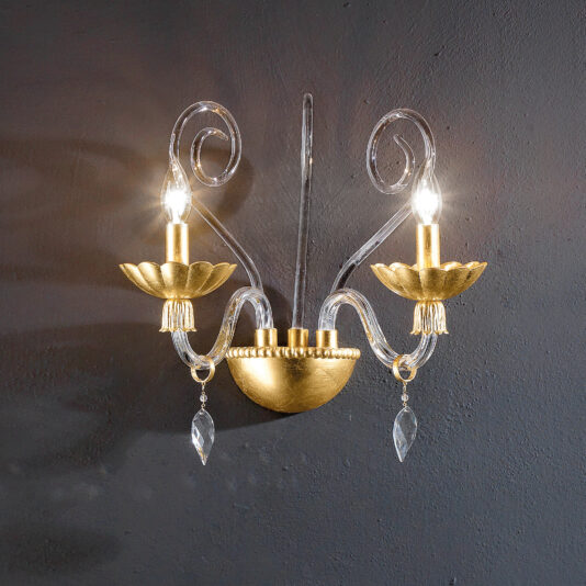 The Modern Double Wall Light With Crystal Drops is mounted on a dark wall, featuring a decorative, classic style with gold accents and translucent elements. Each arm holds a light bulb with a stylized gold holder shaped like flower petals. Crystal pendants hanging from each arm add an elegant touch reminiscent of French design.