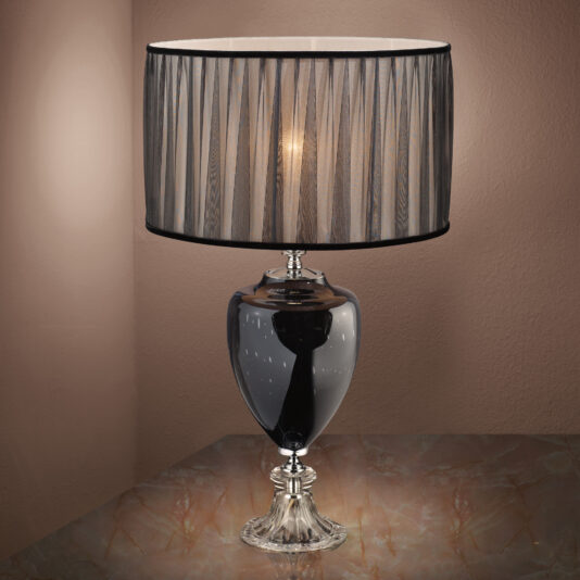 The Modern Glass Table Lamp, featuring an elegant black base and a shiny, translucent finish, stands on a marble surface. It is adorned with a black pleated lampshade that emits a soft, warm light, creating a cozy ambiance against the neutral-colored background wall.