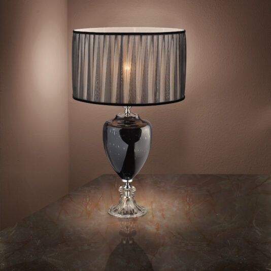 Modern Glass Table Lamp The Modern Glass Table Lamp, featuring a sleek metallic base, stands on a marble surface. Its cylindrical shade, adorned with vertical black stripes, casts a warm, soft glow against a taupe-colored wall.