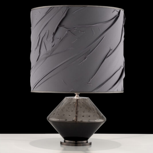 The Modern Hand Blown Glass Table Lamp boasts a textured gray drum lampshade and a striking geometric base with two distinct tones. The glossy black bottom half of the base contrasts beautifully with the translucent upper half, which features an intricate dotted pattern. This elegant lamp is displayed against a black background on a white surface.