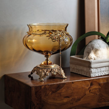 Ornate Jewelled Amber Glass Centrepiece An **Ornate Jewelled Amber Glass Centrepiece** on a decorative gold stand sits on a wooden surface. Beside it is a rectangular planter with a white sculpture of a head, adorned with a bracelet featuring an intricate design. An arched green leaf extends from the planter across the head.