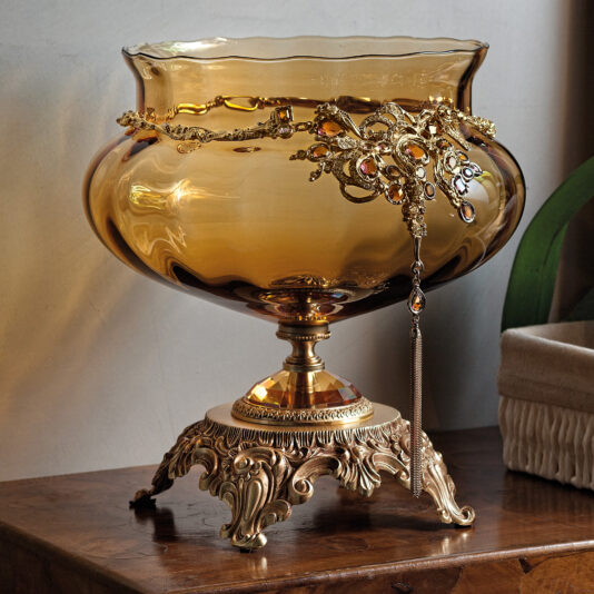 Ornate Jewelled Amber Glass Centrepiece The Ornate Jewelled Amber Glass Centrepiece, featuring an exquisite gold stand and detailed design, is enhanced by a decorative gold chain. It rests on a polished wooden surface, with part of a green leaf and an item draped in white fabric visible in the background.