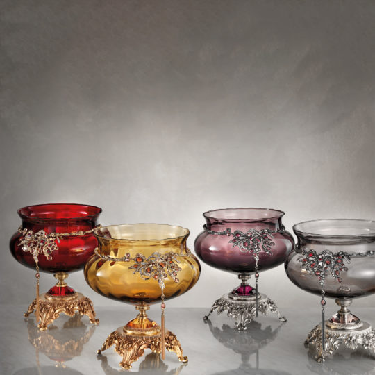 Displayed are four Ornate Jewelled Glass Centrepieces in stunning shades of red, amber, purple, and grey. Each centrepiece boasts decorative metal accents with intricate designs set atop elegant pedestal stands. These luxurious and antique pieces gleam beautifully against a neutral background.