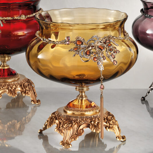Ornate Jewelled Glass Centrepiece The Ornate Jewelled Glass Centrepiece is a decorative, amber-colored glass bowl adorned with ornate, gold-toned metal accents and a hanging tassel. This exquisite piece features an intricate design embellished with gemstones and is showcased on an elaborate stand with curved details. Two similar bowls flank this opulent display.