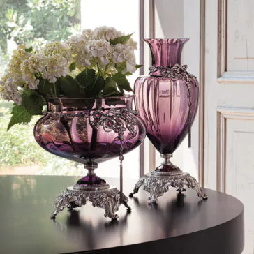 Ornate Jewelled Glass Centrepiece and Vase Two Ornate Jewelled Glass Centrepieces and Vases with intricate silver bases and trim adorn a dark wooden table. One round centrepiece holds a bouquet of white hydrangeas, while the other, tall and fluted, serves as an exquisite centrepiece. Sunlight filters through the window, revealing greenery outside.