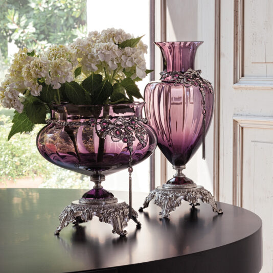 Two Ornate Jewelled Glass Centrepieces and Vases with intricate silver bases and trim adorn a dark wooden table. One round centrepiece holds a bouquet of white hydrangeas, while the other, tall and fluted, serves as an exquisite centrepiece. Sunlight filters through the window, revealing greenery outside.