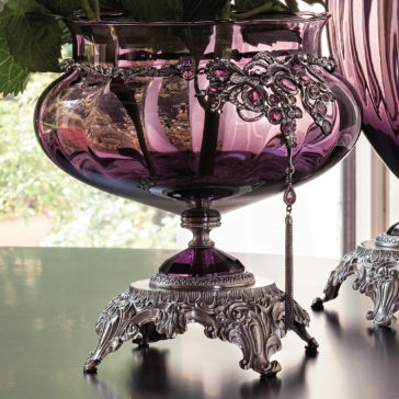 Ornate Jewelled Glass Centrepiece and Vase A close-up of the Ornate Jewelled Glass Centrepiece and Vase reveals its exquisite silver filigree detailing. The vase stands on a decorative silver base adorned with intricate patterns. In the background, light streams through a window, illuminating this stunning piece while offering a softly blurred view of the outdoors.