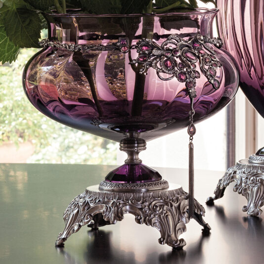 A close-up of the Ornate Jewelled Glass Centrepiece and Vase reveals its exquisite silver filigree detailing. The vase stands on a decorative silver base adorned with intricate patterns. In the background, light streams through a window, illuminating this stunning piece while offering a softly blurred view of the outdoors.