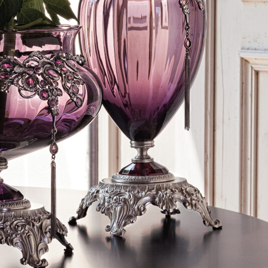 The Ornate Jewelled Glass Centrepiece and Vase features a pair of elegant purple glass vases with intricate silver bases and decorative chains adorned with purple gems. These exquisite centrepieces are showcased on a polished wooden surface against a white wall. Light reflects off the vases, emphasizing their glossy finish.