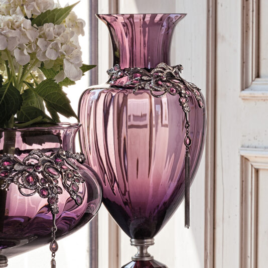 An ornate jewelled glass centrepiece and vase with tall, elegant purple glass featuring silver detailing and dangling jewels stands proudly next to a shorter counterpart, similarly decorated and adorned with white hydrangea flowers. Both pieces are set against a vintage-style wooden background.
