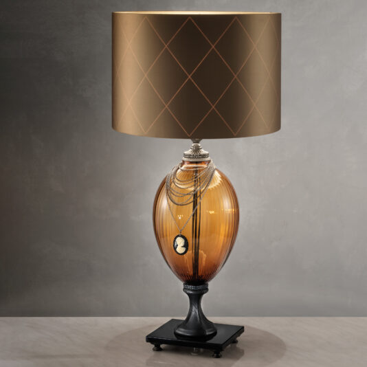The Regency Style Amber Glass Table Lamp features an elegant cameo pendant draped around the top. The beige lampshade showcases a gold crisscross diamond pattern, perfectly complementing its vintage aesthetic. This refined piece stands gracefully on a dark square base against a grey backdrop.