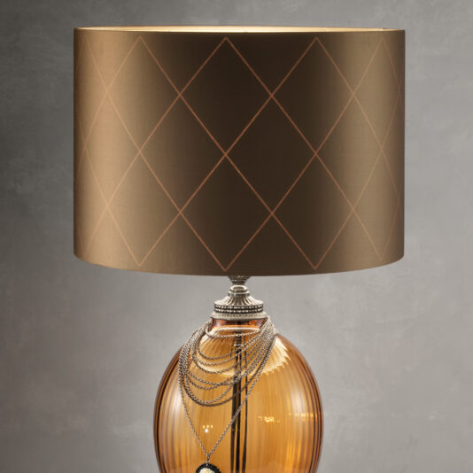 The Regency Style Amber Glass Table Lamp, embellished with decorative silver chains, graces the room with its elegance. Its brown cylindrical shade boasts a subtle geometric diamond pattern, creating a striking contrast against the plain grey wall.
