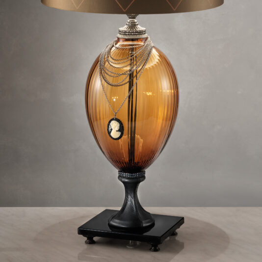 The Regency Style Amber Glass Table Lamp, which has a black pedestal and beige lampshade, graces a light marble surface. Draped over this elegant piece is a layered silver necklace featuring a black and white cameo pendant, all set against a sophisticated gray background.