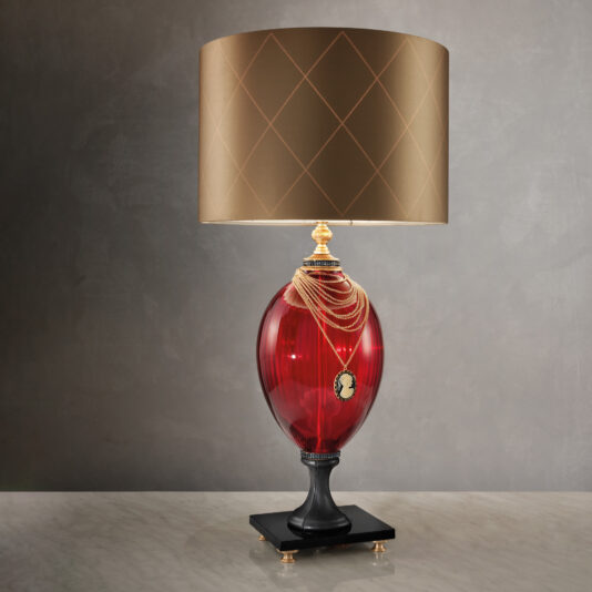 The Regency Style Ruby Glass Table Lamp boasts a substantial, ruby-red oval glass base embellished with an elegant cameo and gold accents. The lamp is topped with a gold-colored lampshade featuring an intricate diamond pattern and is set on a marble surface in front of a neutral backdrop.