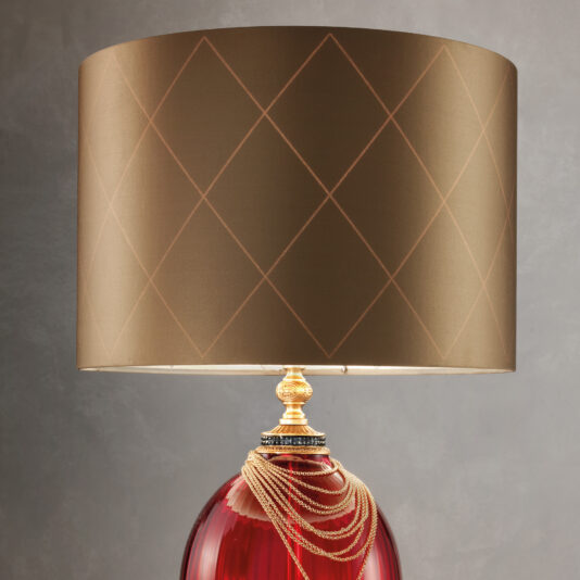 Regency Style Ruby Glass Table Lamp The Regency Style Ruby Glass Table Lamp features a chic design with a large, gold diamond-patterned lampshade and a striking red glass base. Elegantly draped decorative gold chains adorn the top of the base, set against a neutral gray wall.