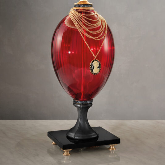 Regency Style Ruby Glass Table Lamp A large, oval-shaped red vase with a ribbed texture is placed on a black pedestal base with four golden feet. Reminiscent of the Regency Style Ruby Glass Table Lamp, it is adorned with a gold necklace draped over the top, featuring a black cameo pendant with a white silhouette profile of a person.