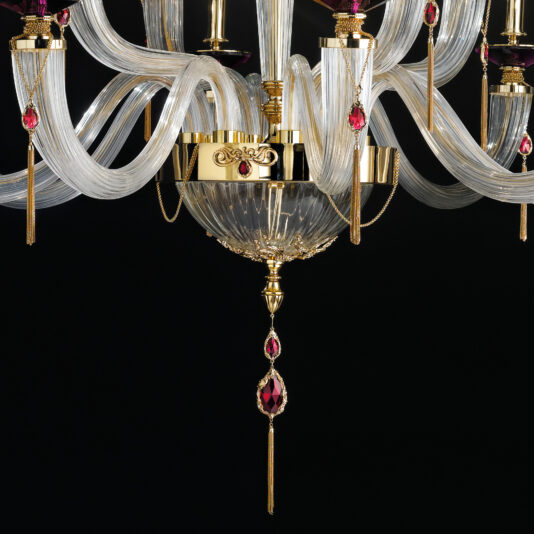 A close-up image of the Swarovski® Crystal Candle Style Chandelier, showcasing its intricate gold and glass details. The chandelier boasts elegant, curved glass arms adorned with gold accents and hanging red crystal ornaments, all set against a black background.
