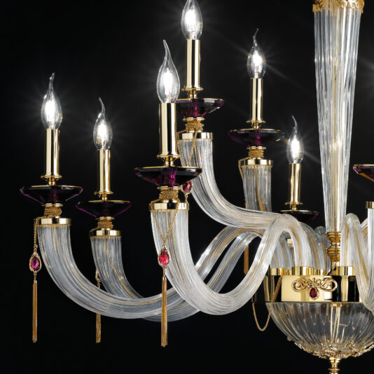 The Swarovski® Crystal Candle Style Chandelier showcases its elegance with multiple curved, transparent arms, each adorned with a decorative light designed to mimic a candle flame. This exquisite chandelier is accented with gold details and hanging tassel ornaments, all set against a dark background.