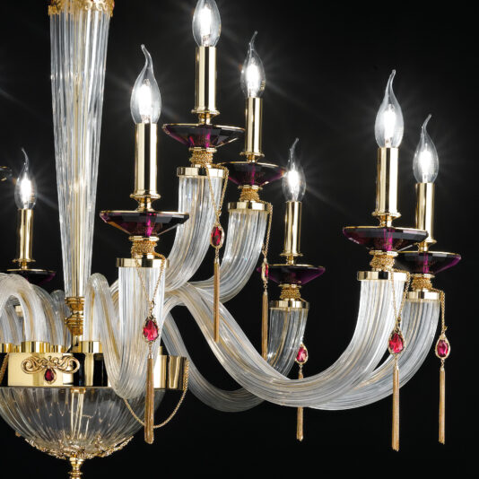 The Swarovski® Crystal Candle Style Chandelier radiates luxury with its gold accents and clear glass arms, embellished with striking red and purple decorative elements. This chandelier boasts multiple candle-shaped light bulbs, casting an elegant glow against a black background.
