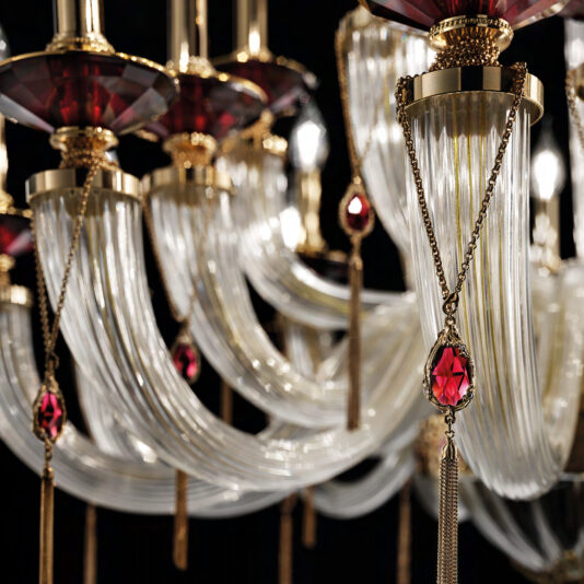 A close-up image of the Swarovski® Crystal Candle Style Chandelier showcases its ornate design with curved glass arms, gold accents, and hanging red crystal pendants. This intricate chandelier reflects light beautifully, highlighting its elegance and luxury.