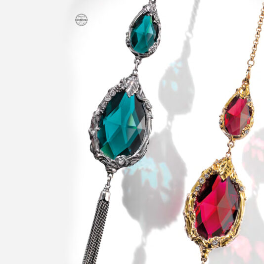 Close-up of two intricate, gemstone-adorned necklaces. The left one features green gemstones framed in silver with a detailed leaf design, reminiscent of the elegance found in a Swarovski® Crystal Candle Style Chandelier. The right one showcases red gemstones set in gold with ornate patterns. Both necklaces reflect light, highlighting their facets.