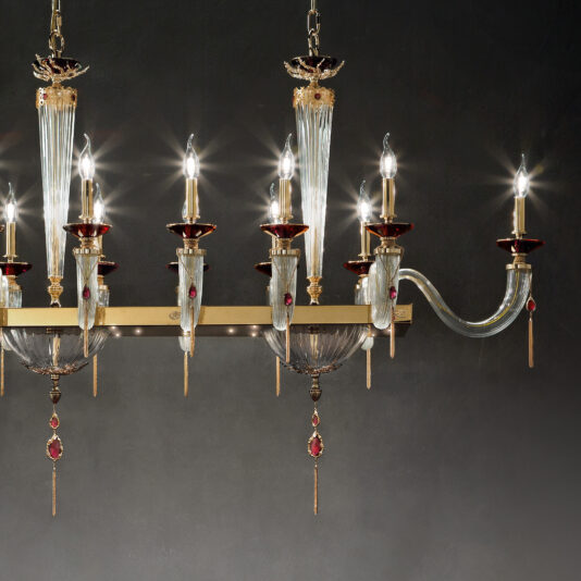 The Swarovski® Crystal Candle Style Snooker Chandelier is a luxurious piece with multiple arms, each adorned with candle-like bulbs made of Swarovski® Crystal. It boasts elegant glass and metal elements embellished with ornate detailing and red accents. The chandelier's intricate design is highlighted against a dark background, making it a stunning centerpiece.