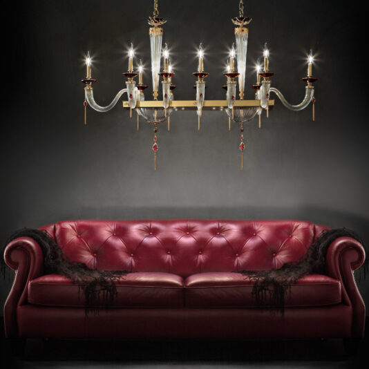 A luxurious red leather tufted sofa with a black lace throw draped over the arm stands against a dark background. Above, an elegant Swarovski® Crystal Candle Style Snooker Chandelier hangs, casting a warm glow over the scene.