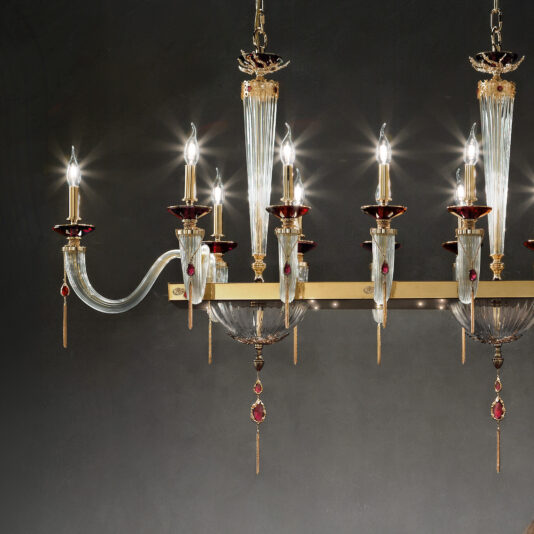 The Swarovski® Crystal Candle Style Snooker Chandelier is an elegant lighting fixture that features multiple candle-like light bulbs. Combining gold, clear glass, and red accent elements with delicate, decorative hanging pendants, this chandelier makes a stunning statement against a dark, neutral wall.