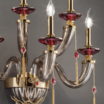 A close-up of an elegant chandelier showcasing gold and crimson accents. The chandelier features multiple curved glass arms adorned with crimson decorative elements and tassels, each capped with candle-like light bulbs reminiscent of a Swarovski® Crystal Candle Style Wall Light. The background is a plain gray wall.