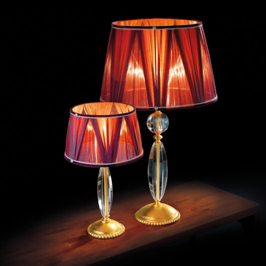 Two Traditional Glass Table Lamps, featuring crystal stems and adorned with red fabric shades, are elegantly placed on a wooden surface against a black background. The larger lamp is positioned slightly behind the smaller one, both emitting a warm glow.