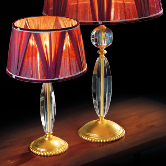 Traditional Glass Table Lamp Two Traditional Glass Table Lamps, featuring elegant golden bases and clear, crystal-like pillars, are topped with rich maroon lampshades and sit on a polished wooden surface. The maroon lampshades emit a warm, soft light that creates a cozy ambiance against the dark background.