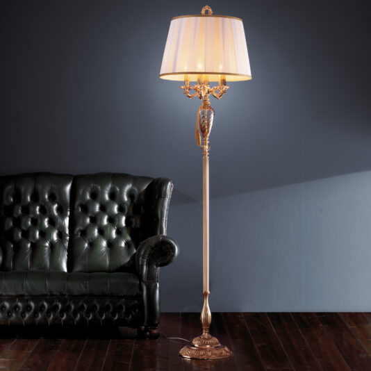 The Candelabra Style Amber Glass Floor Lamp with a gold base and ornate detailing stands beside a dark green, tufted leather sofa. The lamp features a classic white shade and is set against a stark blue-gray wall in a room with dark wooden flooring.