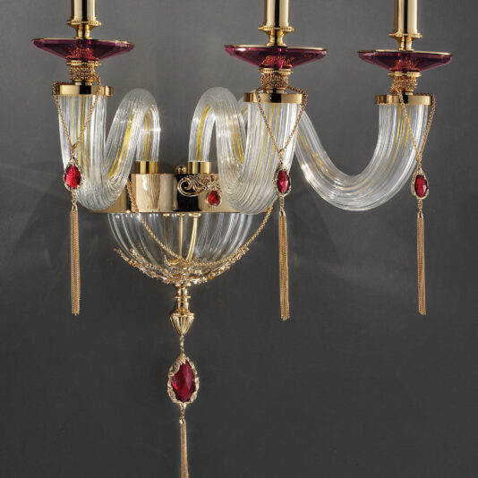 Triple Candle Style Wall Light With Swarovski® Crystals A close-up of the Triple Candle Style Wall Light With Swarovski® Crystals, showcasing gold and crystal accents. The wall light features three candle-style arms adorned with red glass details, hanging tassels, and ornate gold embellishments, all set against a dark gray background. The design exudes sophistication and luxury.