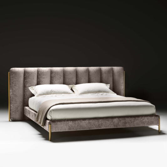 The Contemporary Bed With Wide Headboard boasts a modern design, featuring a wide, tufted headboard upholstered in soft, light gray fabric. The bed is beautifully arranged with neatly placed white linens and pillows, complemented by a beige throw blanket. Its sleek frame is enhanced with golden accents, offering a touch of elegance against the dark background.