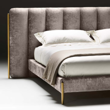 The Contemporary Bed With Wide Headboard is dressed in soft, neutral-toned bedding, including white pillows and a beige blanket. Its plush design is complemented by gold-toned legs that add a touch of elegance against the dark, minimalist backdrop.