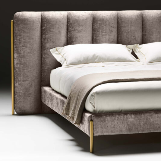 The Contemporary Bed With Wide Headboard is dressed in soft, neutral-toned bedding, including white pillows and a beige blanket. Its plush design is complemented by gold-toned legs that add a touch of elegance against the dark, minimalist backdrop.