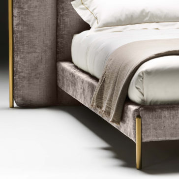 Featured is a close-up of the Contemporary Bed With Wide Headboard, showcasing its soft, gray upholstered frame and gold-colored legs. The bed is elegantly arranged with crisp white sheets and a beige blanket draped over the side. The backdrop is plain and minimalistic.