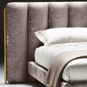 Close-up of the Contemporary Bed With Wide Headboard, showcasing a plush, tufted headboard with a vertical gold accent on the left side. The bed is dressed in white pillows, white bed sheets, and a beige blanket partially folded on top. The headboard fabric appears to be light grey or lavender in color.